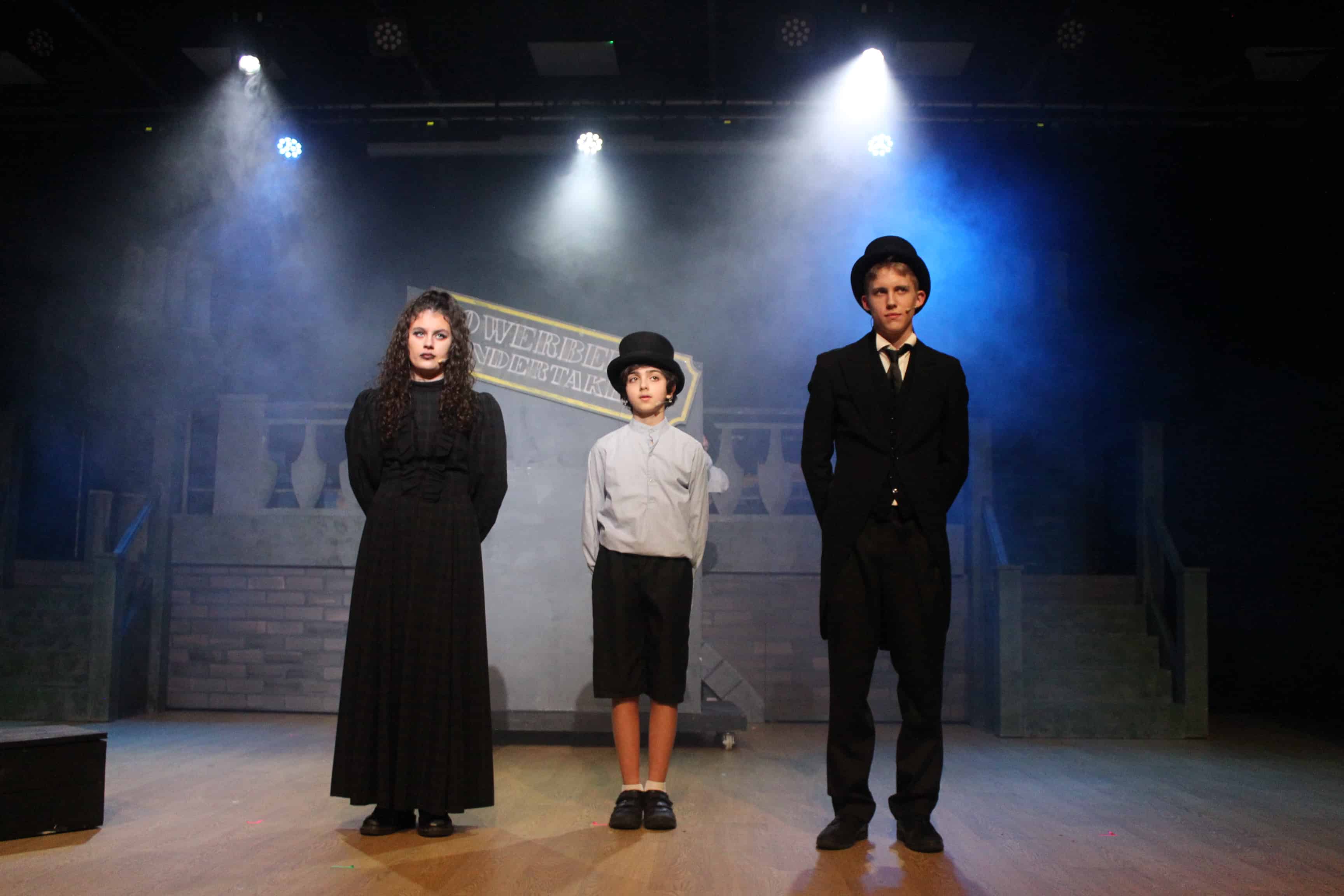 Oliver and Mr and Mrs Sowerberry stand in a line on stage in the Didsbury High School production of Oliver! 2024.