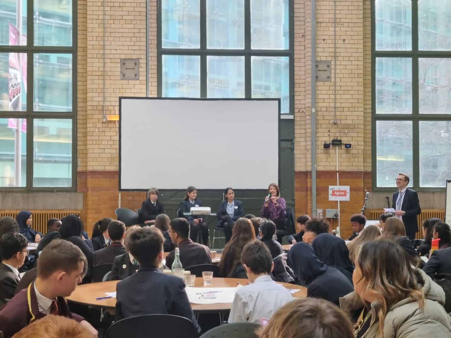 A large crowd of students in the People's History Museum watch students discuss a socio-political issue with a councillor/campaigner